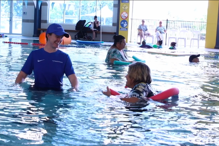 Carol in the pool with her hydrotherapy instructor, both smiling and engaging in exercises with pool noodles