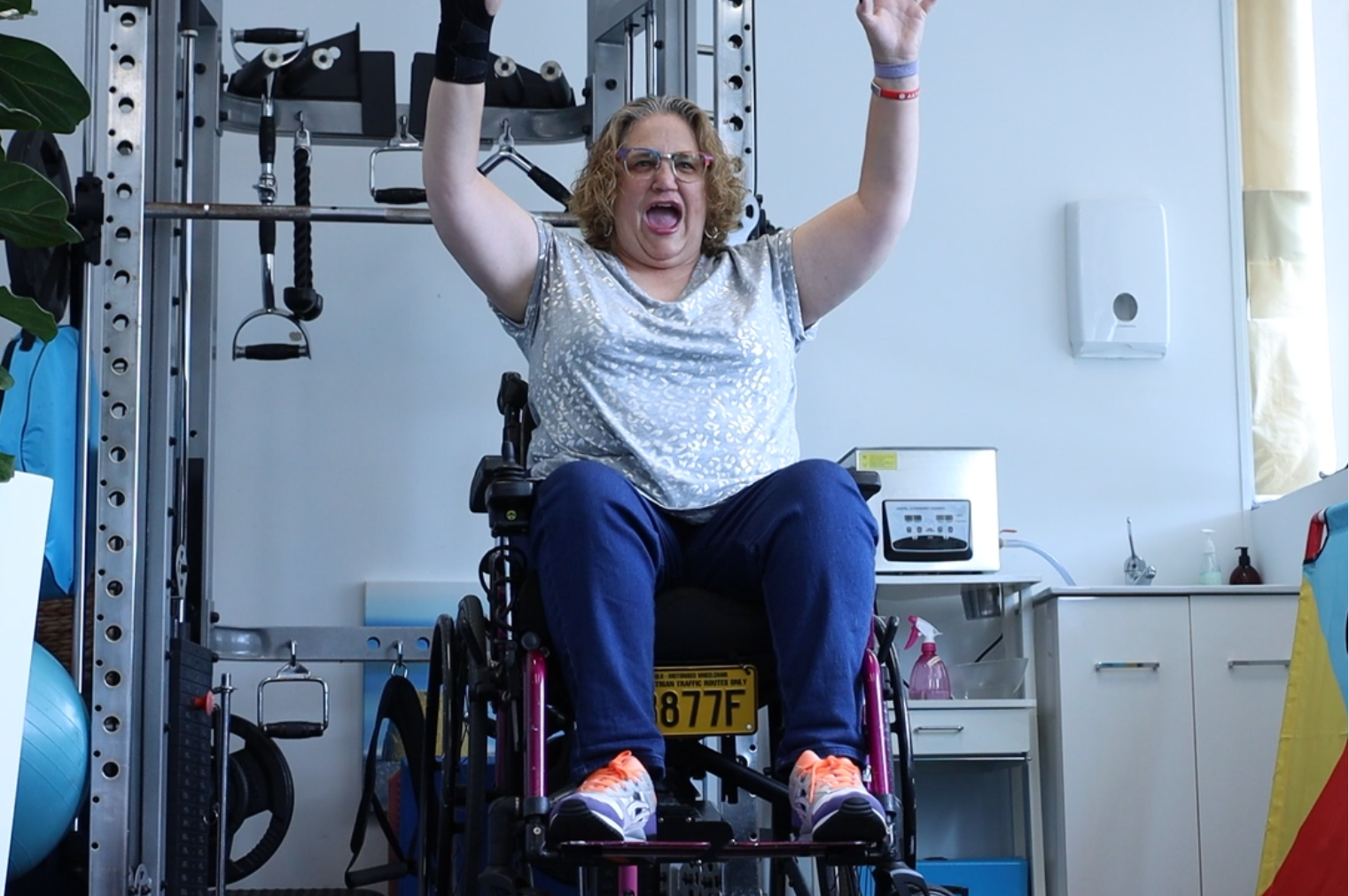 Caroline in a wheelchair raising her arms in celebration inside a gym setting, symbolising her triumph and joy in overcoming challenges with fibromyalgia