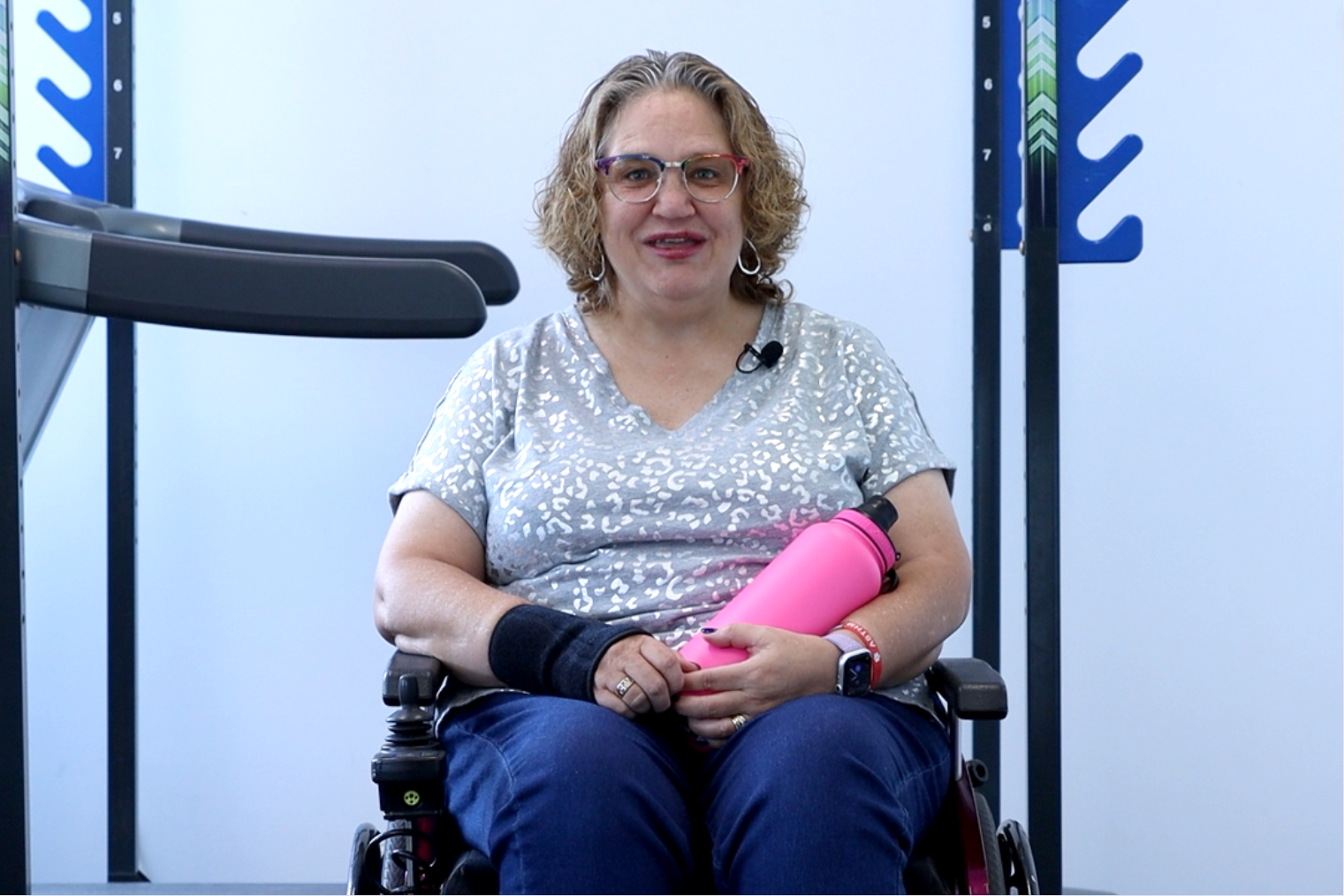 Caroline sitting in her wheelchair holding a pink water bottle, smiling and looking content in a gym environment, showcasing her progress and positive attitude.