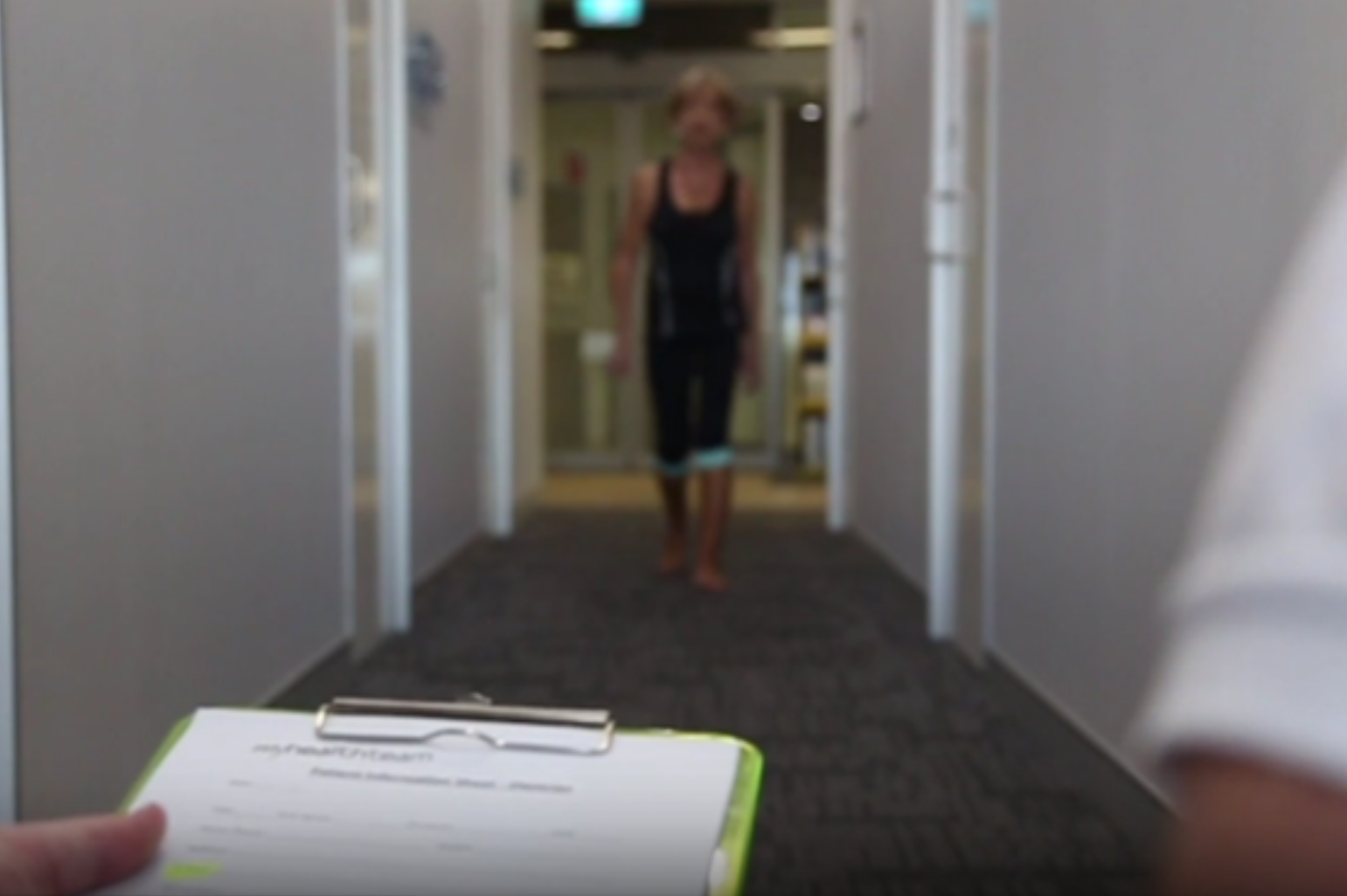 A podiatrist's clipboard is in focus in the foreground, displaying a patient assessment form. In the background, a woman is walking barefoot down a hallway, showcasing her gait. She wears athletic clothing and appears to be participating in a foot assessment.