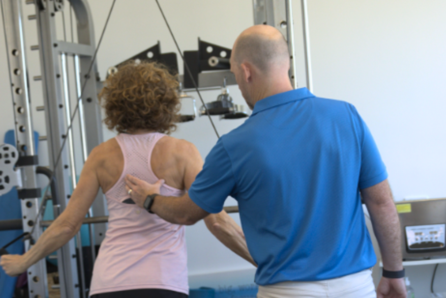 Andrew the director of My Health Team, Redcliffe and an exercise physiologist, assists a woman with curly hair during a workout session, focusing on her shoulder movement.