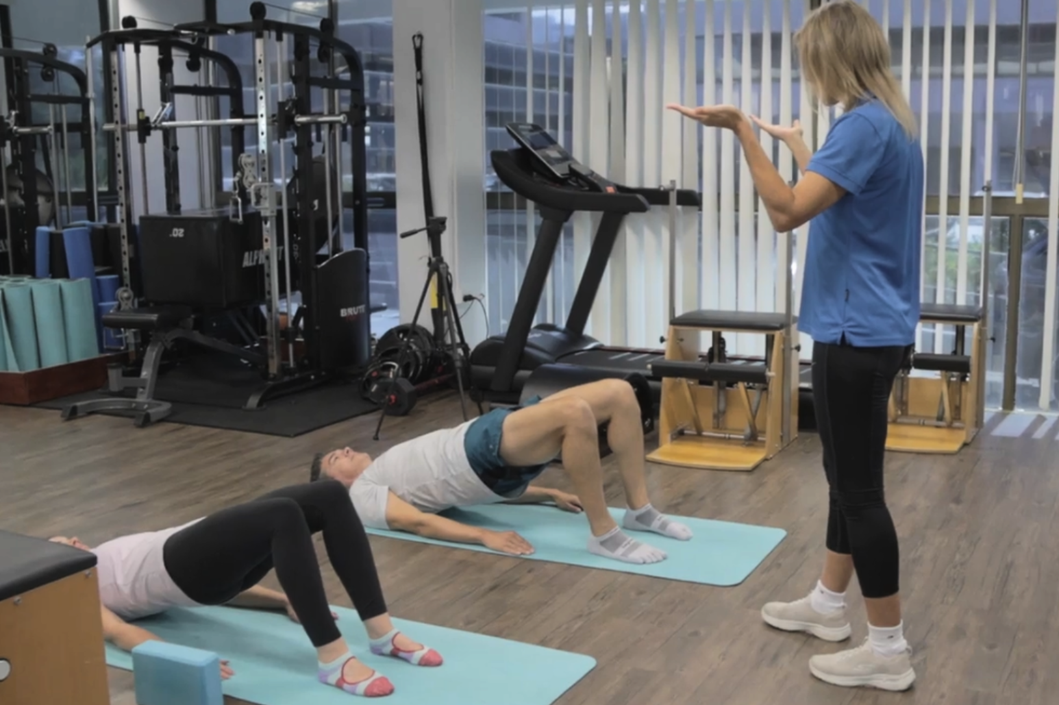 A fitness instructor in a blue shirt demonstrates an exercise to two participants lying on mats in a gym at My Health Team in Redcliffe, where personalised treatment plans are developed for optimal recovery and health.