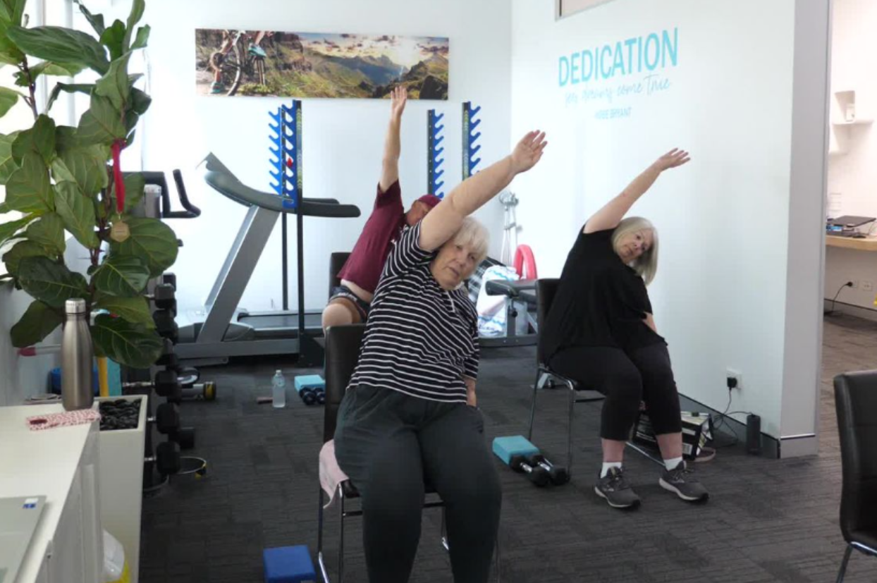 3 elderly people sitting on a chair and engaging in physical therapy at a healthcare facility. The image represents the personalised, supportive care provided by My Health Team in Redcliffe to individuals with chronic diseases and disabilities to improve their health and quality of life.